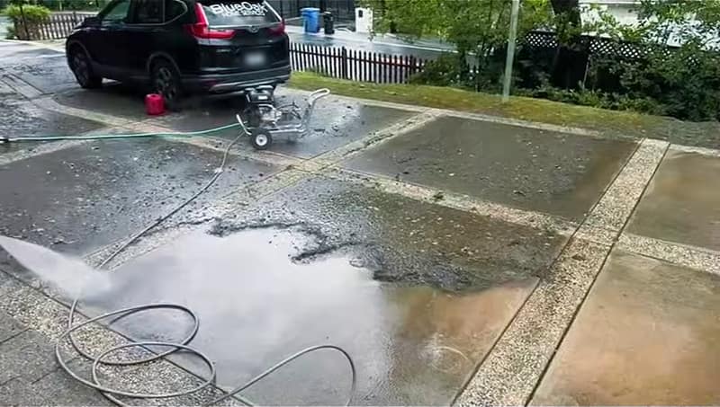 Pressure washer spray clearing mud off a driveway panel, with a gas pressure washer on a cart, a red gas can and our lettered SUV at the top of the driveway