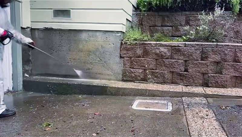 Wand washing a stained concrete foundation wall at the base of a house, beside a block retaining wall