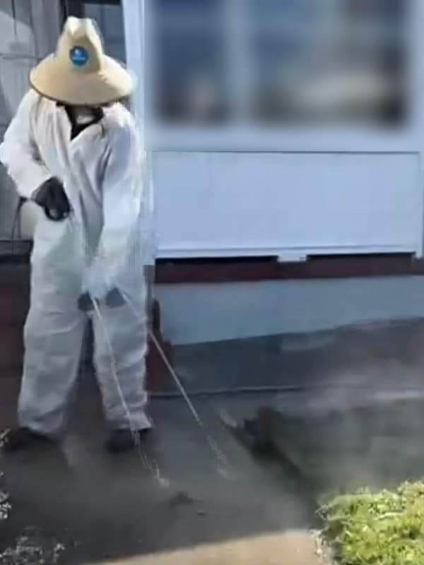 Technician in white coveralls and a straw hat pressure washing a concrete walkway beside a front lawn
