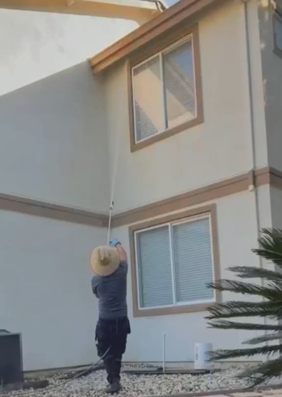 Worker in a straw hat washing the upper wall of a two-story house from the ground with a long wand