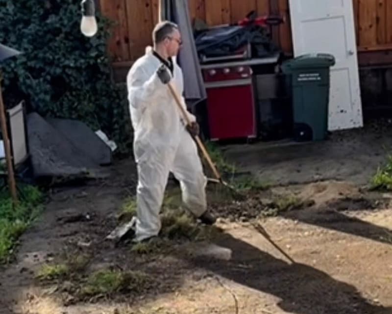 Crew member in white coveralls raking dirt and debris off a backyard concrete slab before pressure washing