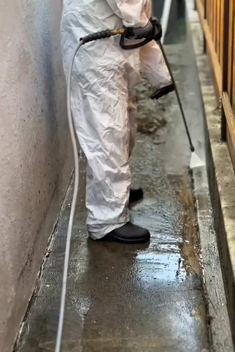 Technician in a white suit pressure washing a narrow side-yard walkway between a house wall and a wood fence