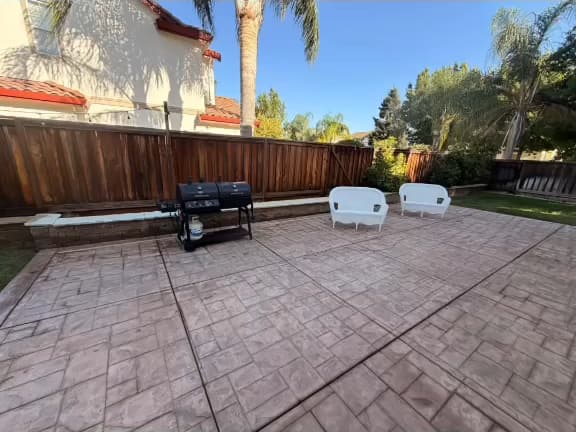 Wide view of a stamped concrete patio after washing, with a grill and two white loveseats by the back fence