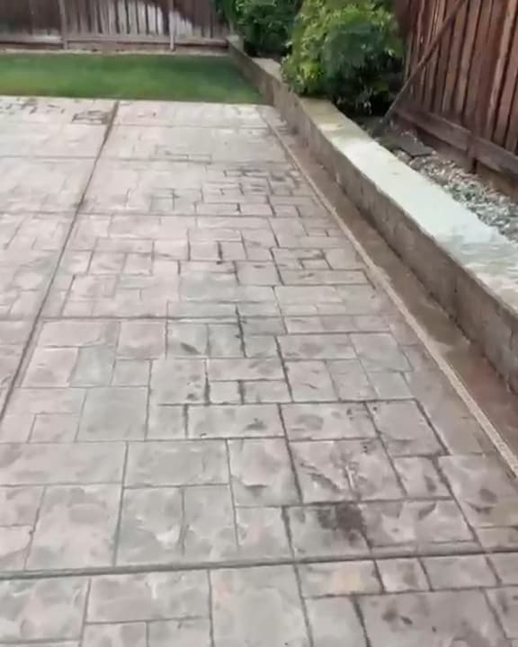 Stamped concrete patio along a block retaining wall, with dark stains and a gray film before washing