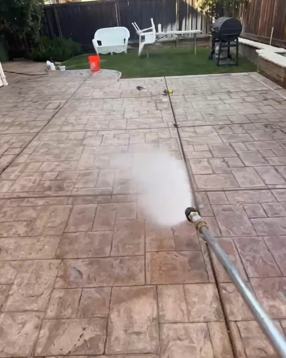 Wand spraying a stamped concrete patio mid-wash, with a lawn, white patio chairs and a grill at the far end